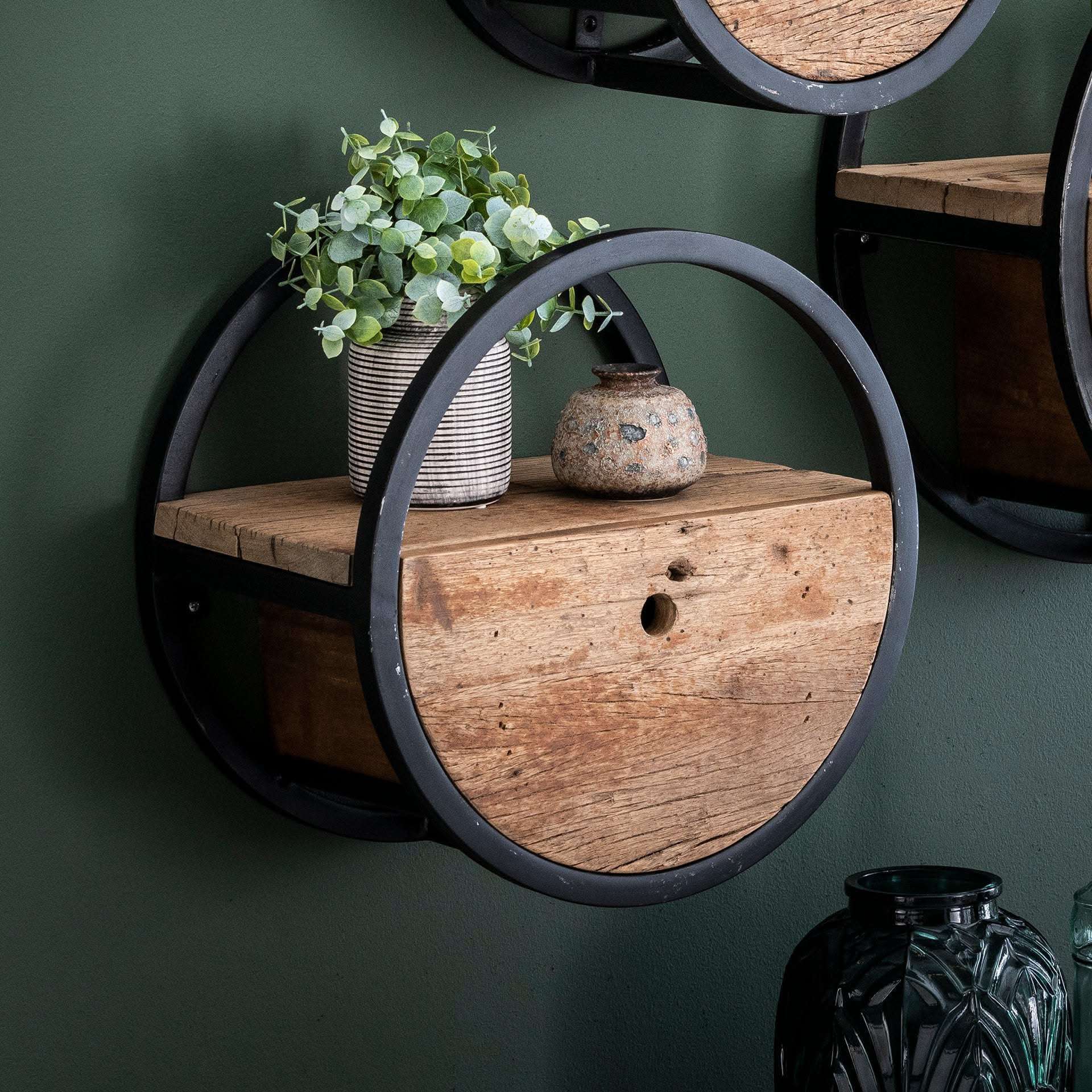 Ein rundes Wandregal mit schwarzem Metallrahmen und Holzboden, darauf eine kleine Pflanze im gestreiften Übertopf und eine Keramikvase.